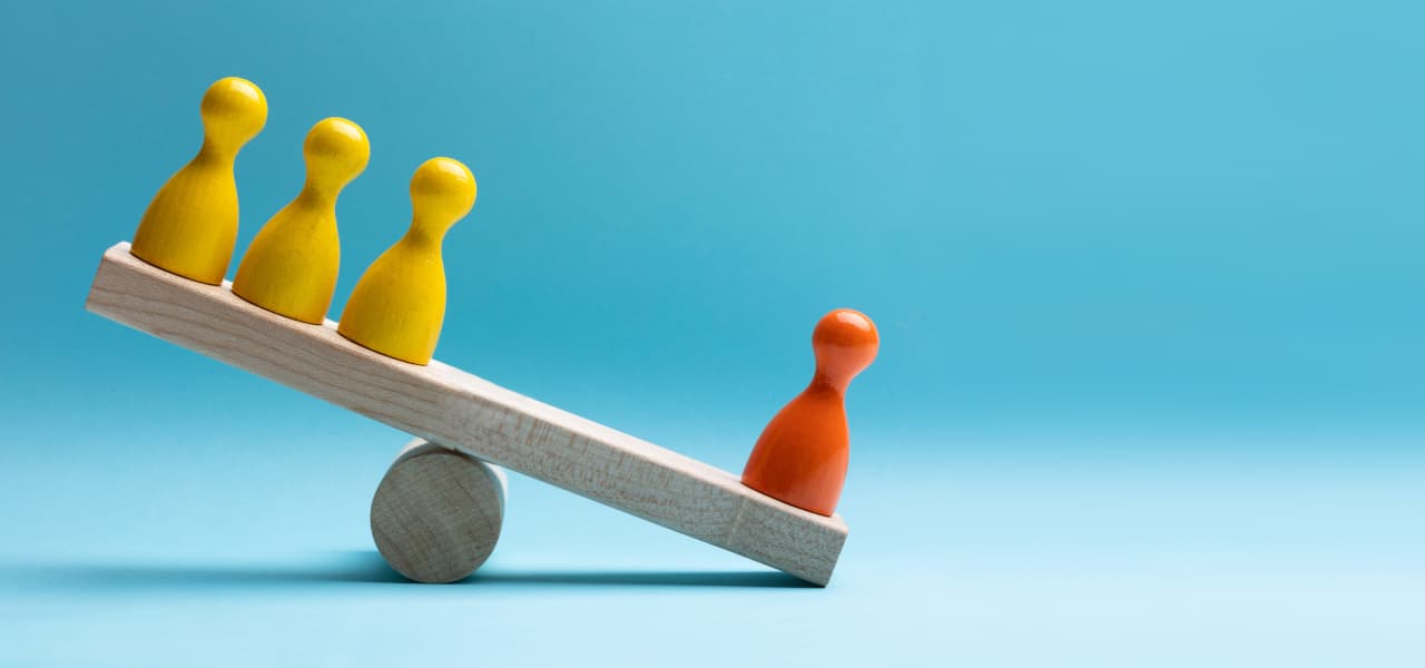 Rote und gelbe Spielfiguren balancieren auf einer hölzernen Wippe | Motiv ©Andrey Popov-stock.adobe.com | © Fotolia.com, April 2026